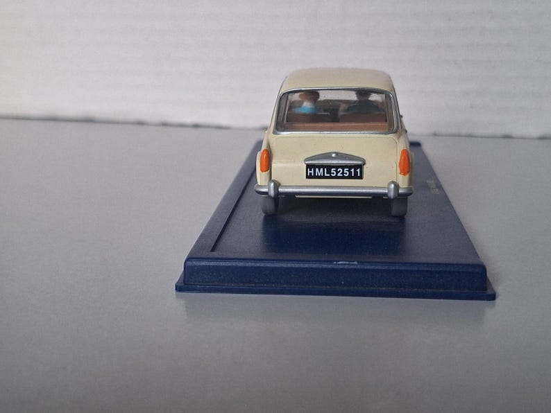 Op de afbeelding: Een cr&egrave;mekleurige miniatuurauto met oranje achterlichten en een zwart kenteken met de tekst "HML52511". De auto staat op een donkerblauwe rechthoekige basis. De ramen van de auto tonen twee figuren binnenin. Een klassiek automodel.
