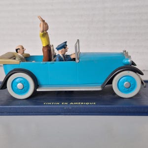 Puede incluir: Figura de coche antiguo azul claro con neum&aacute;ticos de banda blanca y detalles azules. El coche tiene tres figuras, incluida una que saluda. La base dice "TINTIN EN AMERIQUE".