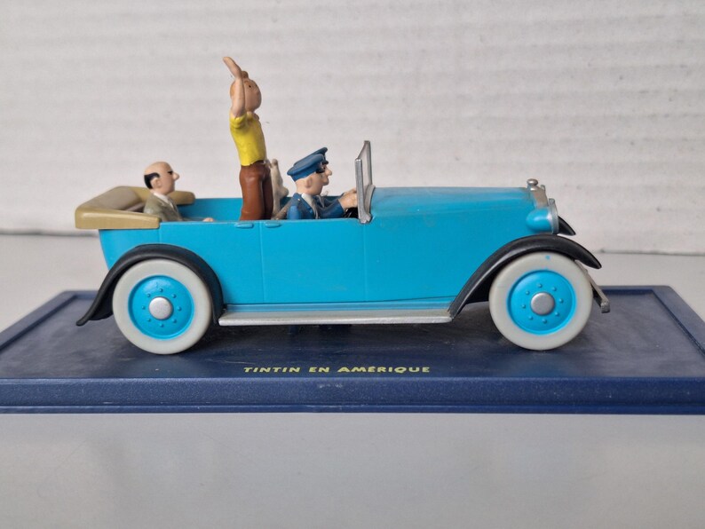 Puede incluir: Figura de coche antiguo azul claro con neum&aacute;ticos de banda blanca y detalles azules. El coche tiene tres figuras, incluida una que saluda. La base dice "TINTIN EN AMERIQUE".