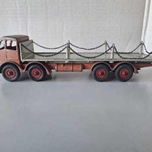 Pode incluir: Um caminhão de brinquedo vintage com um reboque de plataforma e uma cerca de corrente. O caminhão é marrom e branco com rodas vermelhas. O caminhão é feito de metal e tem um design detalhado.