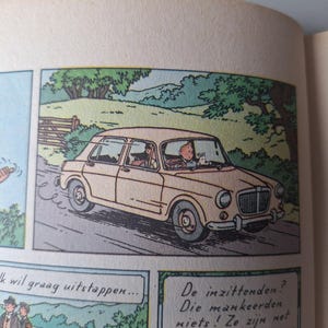 Op de afbeelding: Stripverhaal met een beige auto op een weg. De bestuurder en een passagier zitten in de auto. Een personage in een blauw shirt staat links. Nederlandse tekstballonnen zijn aanwezig.