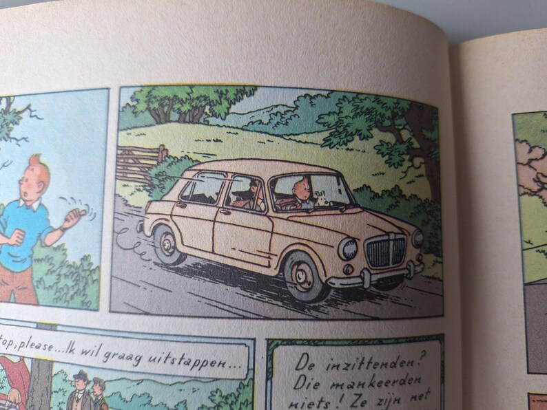 Op de afbeelding: Stripverhaal met een beige auto op een weg. De bestuurder en een passagier zitten in de auto. Een personage in een blauw shirt staat links. Nederlandse tekstballonnen zijn aanwezig.