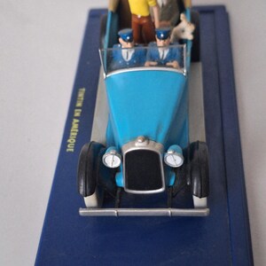 Puede incluir: Un coche de juguete azul con neum&aacute;ticos negros y detalles plateados, con figuras en miniatura de personajes. El coche tiene parabrisas y faros. La base es azul y se ve el texto "TINTIN EN AMERIQUE".