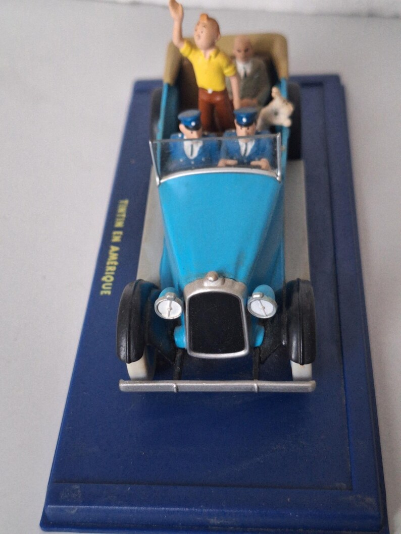 Puede incluir: Un coche de juguete azul con neum&aacute;ticos negros y detalles plateados, con figuras en miniatura de personajes. El coche tiene parabrisas y faros. La base es azul y se ve el texto "TINTIN EN AMERIQUE".
