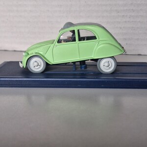 May include: A light green model car with a gray roof and wheels, displayed on a dark blue rectangular base. The car has a classic design, with a rounded body and visible windows. The model car is a collectible item.