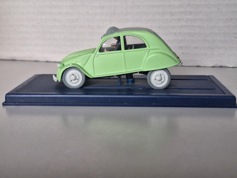 May include: A light green model car with a gray roof and wheels, displayed on a dark blue rectangular base. The car has a classic design, with a rounded body and visible windows. The model car is a collectible item.