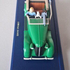 May include: A green vintage car model with a tan roof and brown interior. A small figure of a person in a blue shirt is driving, with a white dog in the passenger seat. The model sits on a blue base with the text "L'OREILLE CASSEE".
