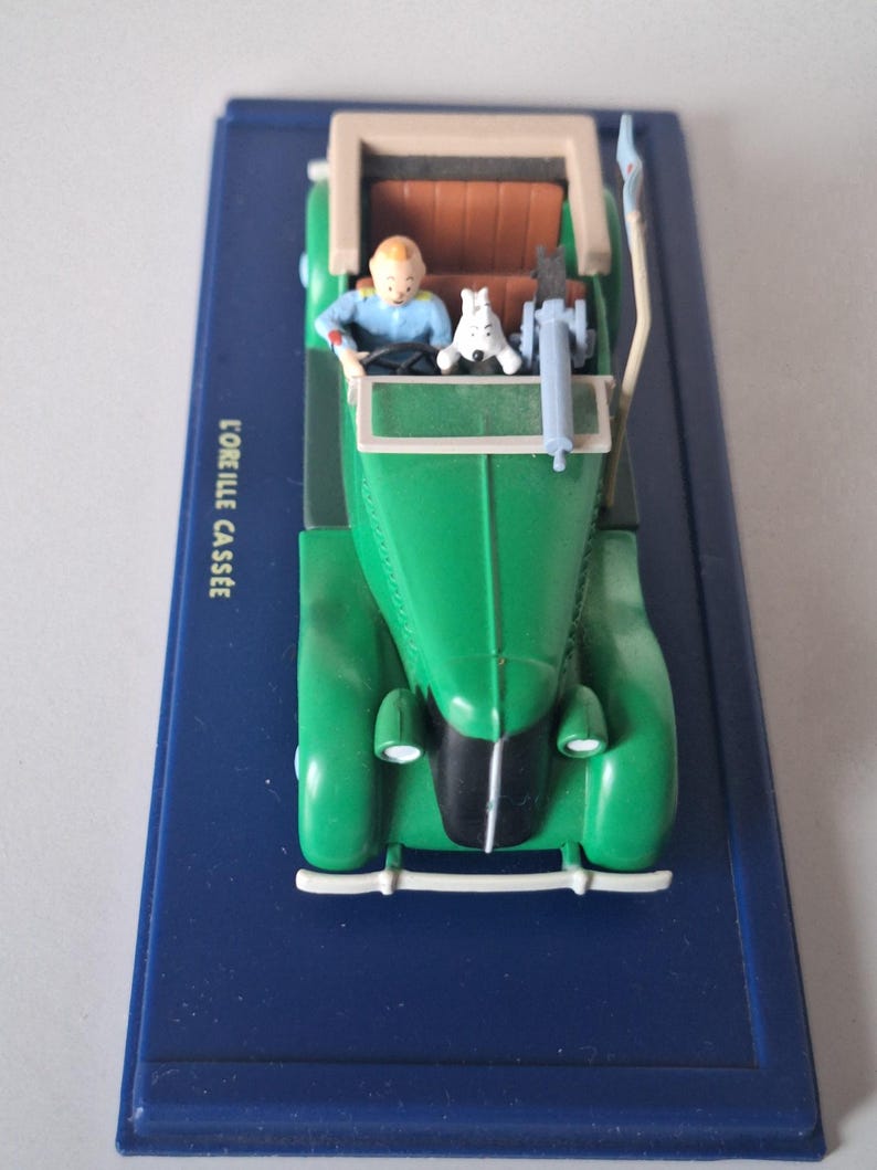 May include: A green vintage car model with a tan roof and brown interior. A small figure of a person in a blue shirt is driving, with a white dog in the passenger seat. The model sits on a blue base with the text "L'OREILLE CASSEE".