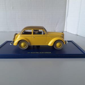 May include: A yellow vintage car model with a brown roof, displayed on a dark blue rectangular base. The car has a classic design with rounded fenders and a detailed interior. The text "LE SCEPTRE D'OTTOKAR" is printed on the base.
