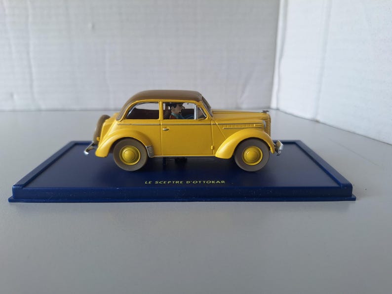 May include: A yellow vintage car model with a brown roof, displayed on a dark blue rectangular base. The car has a classic design with rounded fenders and a detailed interior. The text "LE SCEPTRE D'OTTOKAR" is printed on the base.