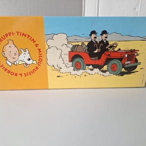 May include: A rectangular box featuring illustrations from the Tintin comic series. The left side has a portrait of Tintin and Snowy, with text in a circle. The right side depicts a red jeep driving through a desert.