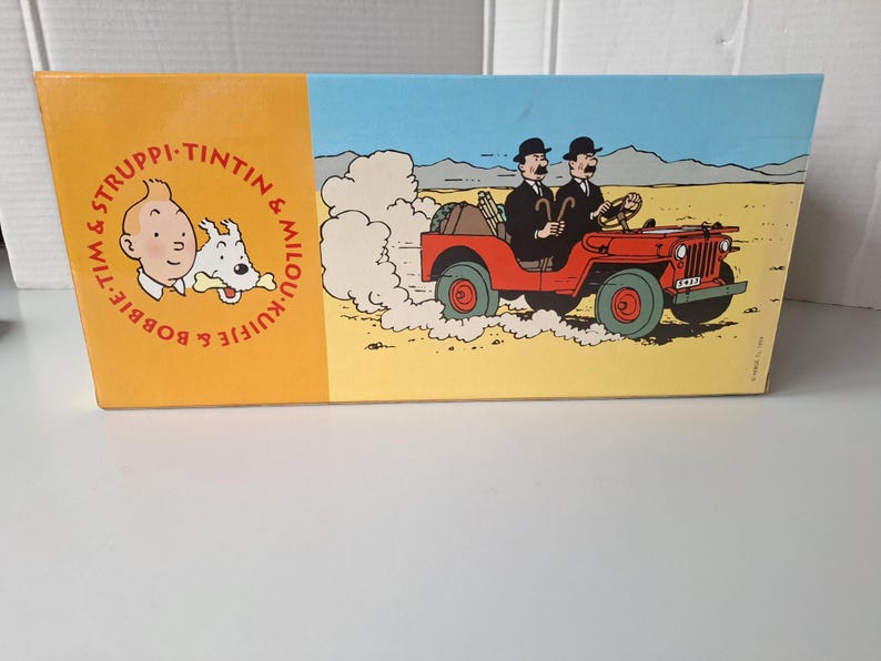 May include: A rectangular box featuring illustrations from the Tintin comic series. The left side has a portrait of Tintin and Snowy, with text in a circle. The right side depicts a red jeep driving through a desert.