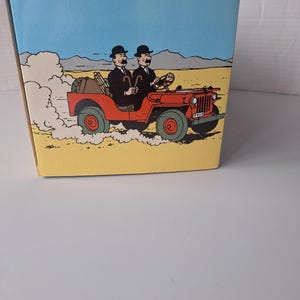 May include: A box featuring a cartoon illustration of two men in black suits and bowler hats driving a red jeep through a desert landscape. The jeep has green wheels and a license plate.