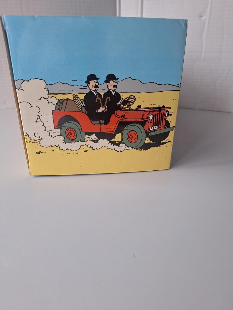 May include: A box featuring a cartoon illustration of two men in black suits and bowler hats driving a red jeep through a desert landscape. The jeep has green wheels and a license plate.