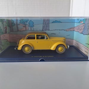 May include: A yellow vintage car model with a brown roof, displayed in a clear plastic case. The car is set against a backdrop of a cartoon landscape. The text "LE SCEPTRE D'OTTOKAR" is visible on the base.