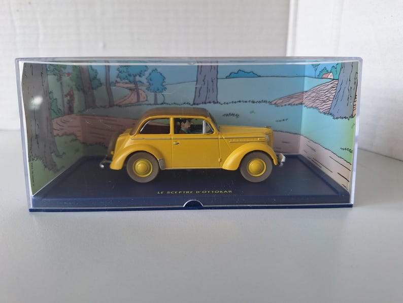 May include: A yellow vintage car model with a brown roof, displayed in a clear plastic case. The car is set against a backdrop of a cartoon landscape. The text "LE SCEPTRE D'OTTOKAR" is visible on the base.