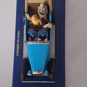 Puede incluir: Un coche de juguete azul con figuras de personajes de la serie de c&oacute;mics Tint&iacute;n. El coche tiene una carrocer&iacute;a azul claro, ruedas negras y un interior beige. Las figuras incluyen a Tint&iacute;n, Mil&uacute; y dos polic&iacute;as. La base es azul oscuro, con el texto "TINTIN EN AMERIQUE".