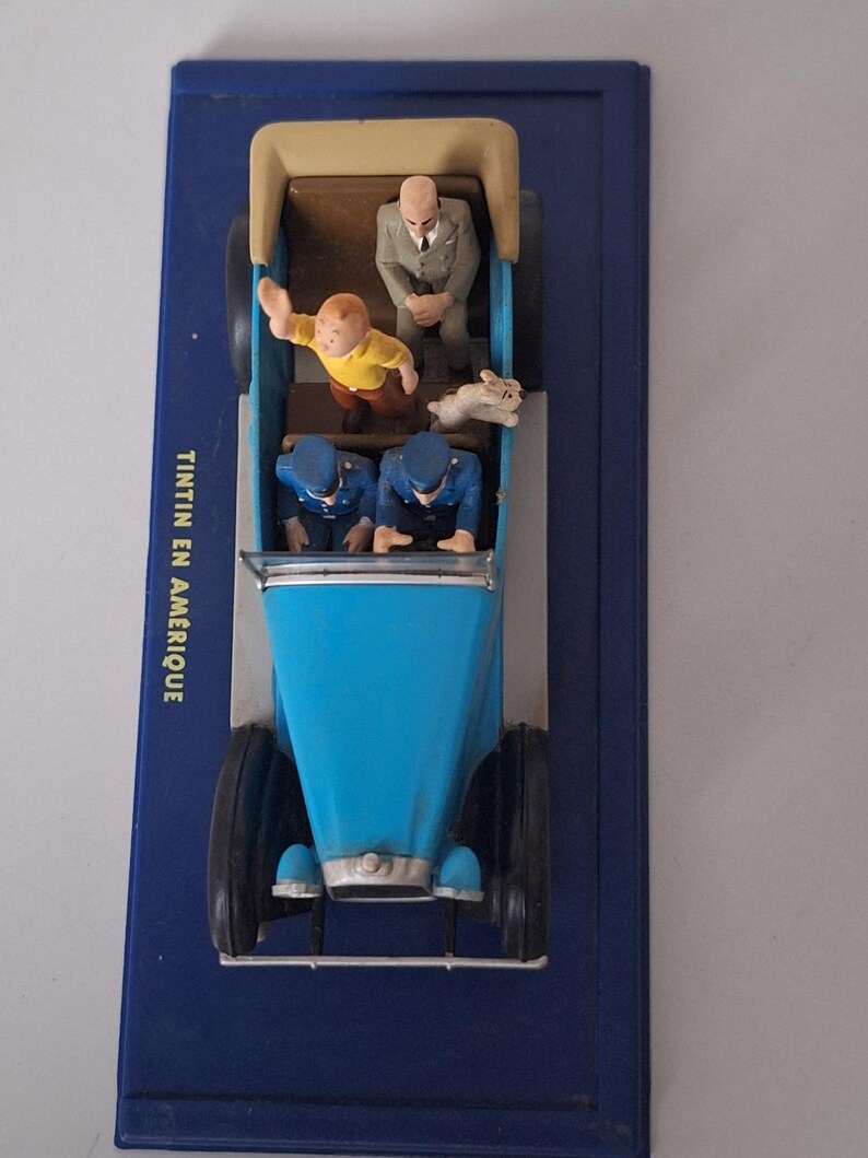 Puede incluir: Un coche de juguete azul con figuras de personajes de la serie de c&oacute;mics Tint&iacute;n. El coche tiene una carrocer&iacute;a azul claro, ruedas negras y un interior beige. Las figuras incluyen a Tint&iacute;n, Mil&uacute; y dos polic&iacute;as. La base es azul oscuro, con el texto "TINTIN EN AMERIQUE".