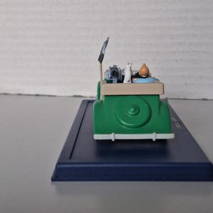 May include: A green toy car with a white front bumper and a beige top. The car features a figure of a person with an orange head and a white cat. The car is on a blue base.