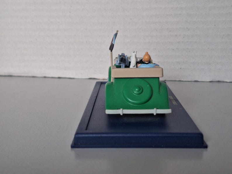 May include: A green toy car with a white front bumper and a beige top. The car features a figure of a person with an orange head and a white cat. The car is on a blue base.