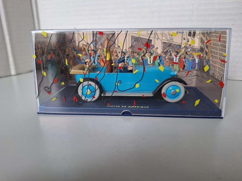Puede incluir: Una maqueta de un coche antiguo azul claro con neum&aacute;ticos blancos, con personajes en el interior, sobre un fondo de multitud y confeti. El coche est&aacute; en una vitrina transparente con el texto "TINTIN EN AMERIQUE" en la base.