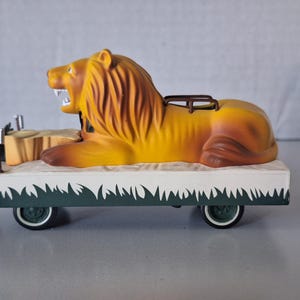 May include: A vintage toy lion figure with an open mouth, lying on a white and green decorated cart. The lion is yellow and brown, with a brown mane. The cart has dark green wheels.