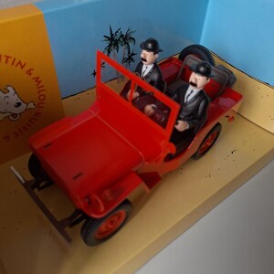 May include: A red toy jeep with two figures wearing black suits and bowler hats. The jeep is on a diorama with a beach and palm tree backdrop. The figures resemble characters from a comic book.