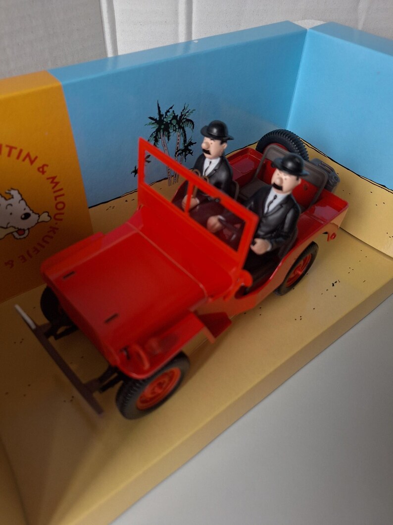May include: A red toy jeep with two figures wearing black suits and bowler hats. The jeep is on a diorama with a beach and palm tree backdrop. The figures resemble characters from a comic book.