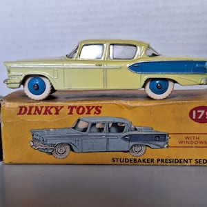 Könnte beinhalten: Ein Dinky Toys Studebaker President Sedan Modellauto aus Metall in Hellgelb mit blauem Heckteil und blauen Rädern mit weißen Reifen. Das Auto steht auf seiner originalen gelben Pappschachtel mit der Aufschrift Dinky Toys 179 with windows.