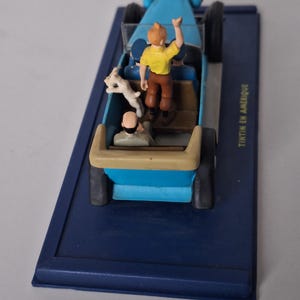 Puede incluir: Un coche de juguete azul con figuras de Tint&iacute;n, Mil&uacute; y otro personaje. El coche est&aacute; sobre una base azul con el texto "Tint&iacute;n en Am&eacute;rique". Tint&iacute;n saluda desde el coche.