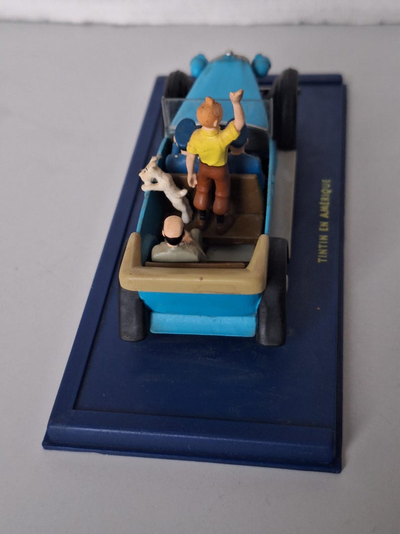 Puede incluir: Un coche de juguete azul con figuras de Tint&iacute;n, Mil&uacute; y otro personaje. El coche est&aacute; sobre una base azul con el texto "Tint&iacute;n en Am&eacute;rique". Tint&iacute;n saluda desde el coche.