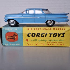Könnte beinhalten: Ein hellblaues Druckguss-Modellauto mit silbernen Rädern steht auf einer Vintage-Corgi-Toys-Box. Die Box ist gelb und blau und zeigt "CORGI TOYS" und "with spring suspension". Das Auto ist ein klassisches Modell.