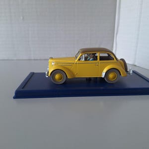 May include: A yellow vintage model car with a brown roof, parked on a dark blue rectangular base. The car has a spare tire on the back and is a classic design. The model car is a collectible item.