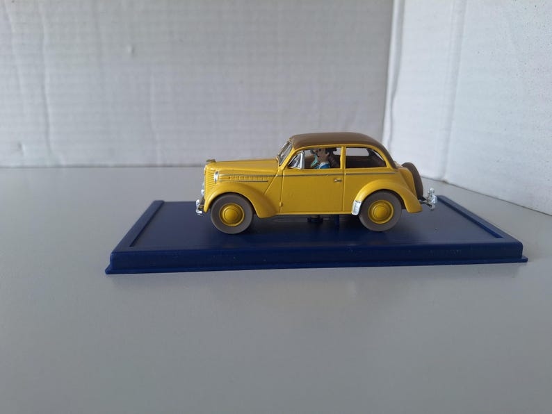 May include: A yellow vintage model car with a brown roof, parked on a dark blue rectangular base. The car has a spare tire on the back and is a classic design. The model car is a collectible item.