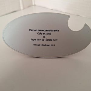 May include: A light grey, oval-shaped object with a curved cutout. Text reads "L'avion de reconnaissance Coke en stock Pages 51 et 52 - &Eacute;chelle 1:72&deg; &copy; Herg&eacute; - Moulinsart 2014". The object is on a white surface.