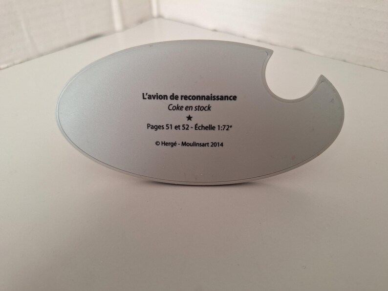 May include: A light grey, oval-shaped object with a curved cutout. Text reads "L'avion de reconnaissance Coke en stock Pages 51 et 52 - &Eacute;chelle 1:72&deg; &copy; Herg&eacute; - Moulinsart 2014". The object is on a white surface.