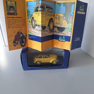 May include: A yellow die-cast model car in a blue box, with a folded comic book featuring a yellow car and the text "TINTIN" in the background. The comic book also shows a motorcycle and text in Dutch.