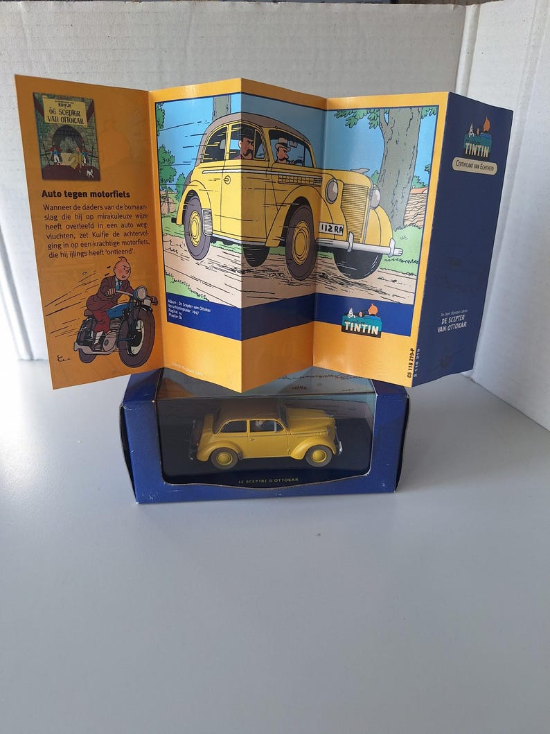 May include: A yellow die-cast model car in a blue box, with a folded comic book featuring a yellow car and the text "TINTIN" in the background. The comic book also shows a motorcycle and text in Dutch.
