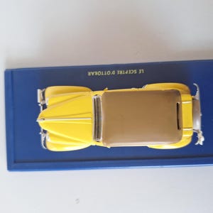 May include: A yellow vintage car model with a brown roof sits on a blue rectangular base. The model has silver accents and is a detailed replica of a classic automobile. The base has text in white.