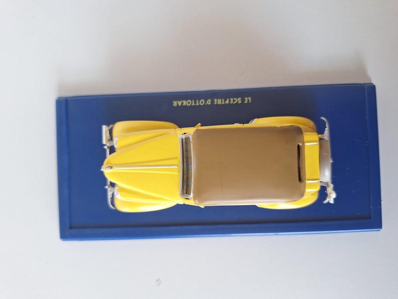 May include: A yellow vintage car model with a brown roof sits on a blue rectangular base. The model has silver accents and is a detailed replica of a classic automobile. The base has text in white.