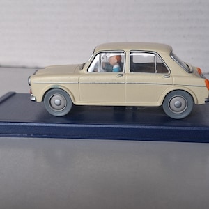 Op de afbeelding: Een beige vintage modelauto met grijze wielen en een blauwe basis. De auto heeft een bestuurdersfiguur binnenin. Het ontwerp van de auto doet denken aan klassieke auto's. Het model is een verzamelobject.