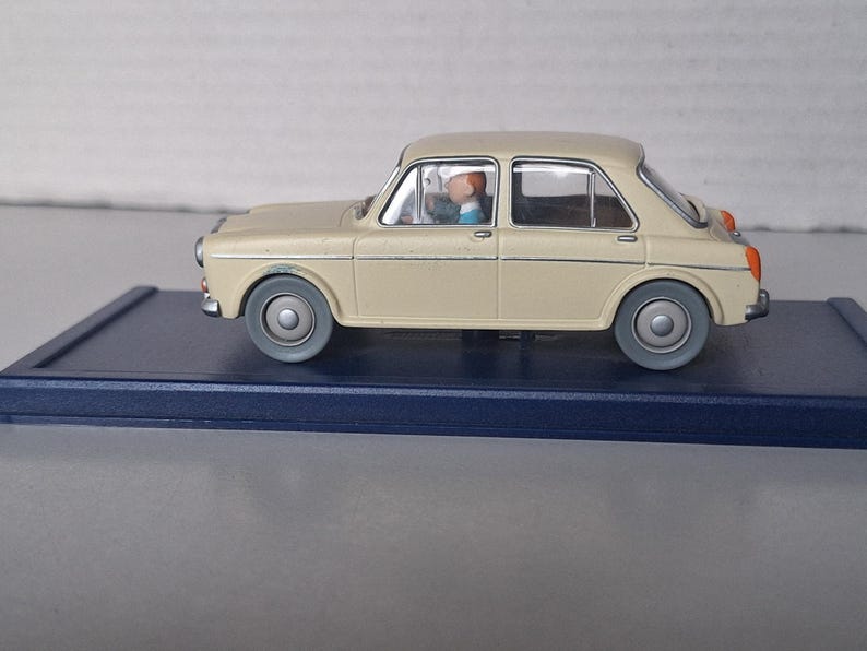 Op de afbeelding: Een beige vintage modelauto met grijze wielen en een blauwe basis. De auto heeft een bestuurdersfiguur binnenin. Het ontwerp van de auto doet denken aan klassieke auto's. Het model is een verzamelobject.