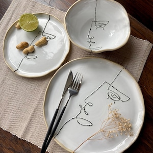 May include: Three white ceramic plates with black line drawings of faces. The plates are on a beige placemat on a wooden table.