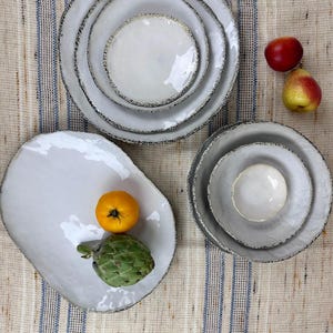 May include: A selection of white ceramic plates and bowls with textured, dark grey rims. An oblong plate holds a yellow tomato and a green artichoke. Two pieces of fruit are also present.