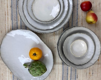Puede incluir: Una colección de platos y cuencos de cerámica blanca con bordes texturizados de color gris oscuro. Un plato oblongo contiene un tomate amarillo y una alcachofa verde. También hay dos frutas presentes.