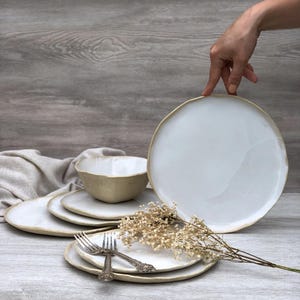 Op de afbeelding: Set witte keramische dinerborden met een licht verhoogde rand. De borden zijn op elkaar gestapeld, met een kom en twee vorken erop. De borden hebben een rustieke, handgemaakte uitstraling.
