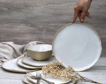 May include: Set of white ceramic dinner plates with a slightly raised rim. The plates are stacked on top of each other, with a bowl and two forks on top. The plates have a rustic, handmade look.
