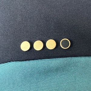 Op de afbeelding: Vijf ronde gouden knoppen met een zwarte knop in het midden. De knoppen zijn in een rij op een donkerblauwe stoffen achtergrond gerangschikt.