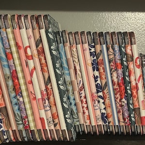 May include: A collection of rectangular tablet cases arranged in a row, each featuring distinct patterns including floral prints, checkered designs, and solid colors in shades of pink, blue, green, and white, with visible camera cutouts on the top.