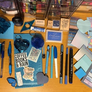 Puede incluir: Una colección de artículos de papelería azules y blancos, como bolígrafos, marcadores, pegatinas y sellos, dispuestos sobre una superficie de madera. Se ve un contenedor de almacenamiento transparente en el fondo. Algunos artículos tienen texto.